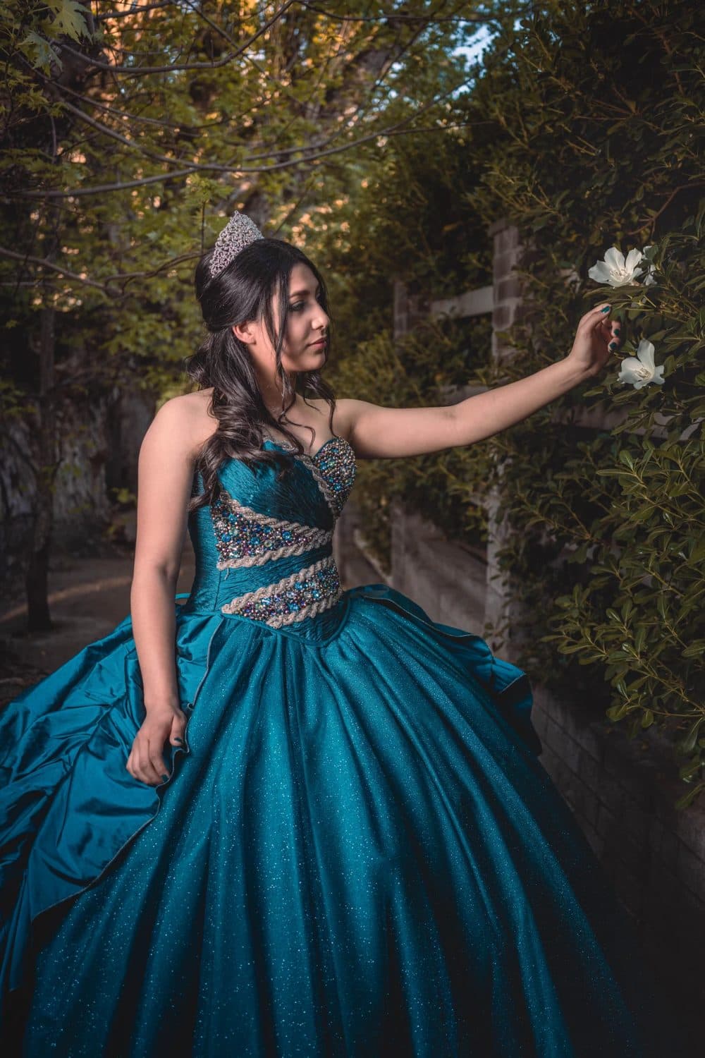 Camila con vestido verde azulado y tiara, tocando una flor blanca en un jardín